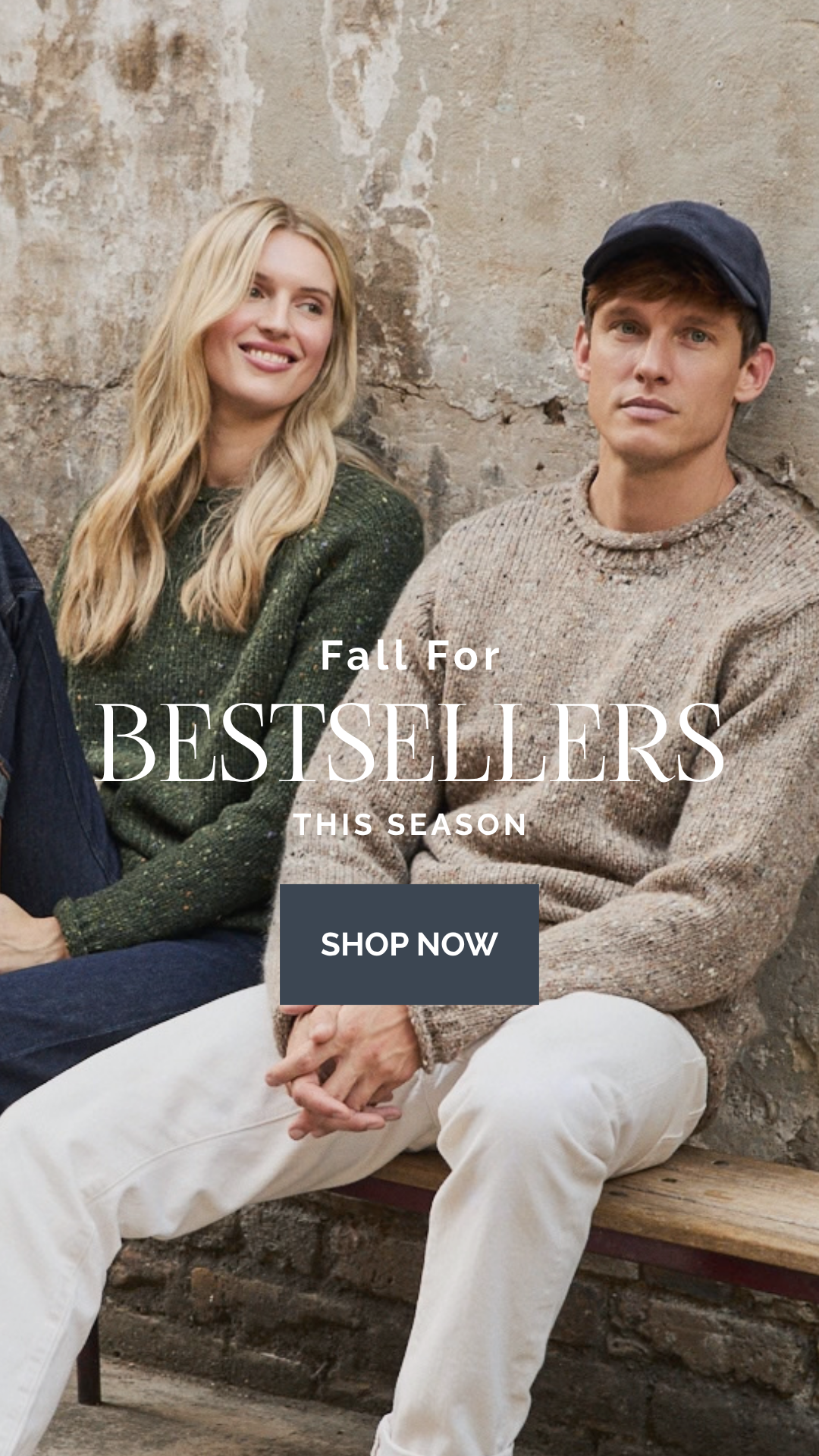 A man and a woman sitting on a bench against a rustic stone wall, wearing cozy knit sweaters and casual clothing. Large overlaid text reads 'Fall For BESTSELLERS THIS SEASON' with a 'SHOP NOW' button.